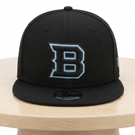 A black flat-brim baseball cap with a large outlined 'B' logo on the front, displayed on a light wood round table against a white background.