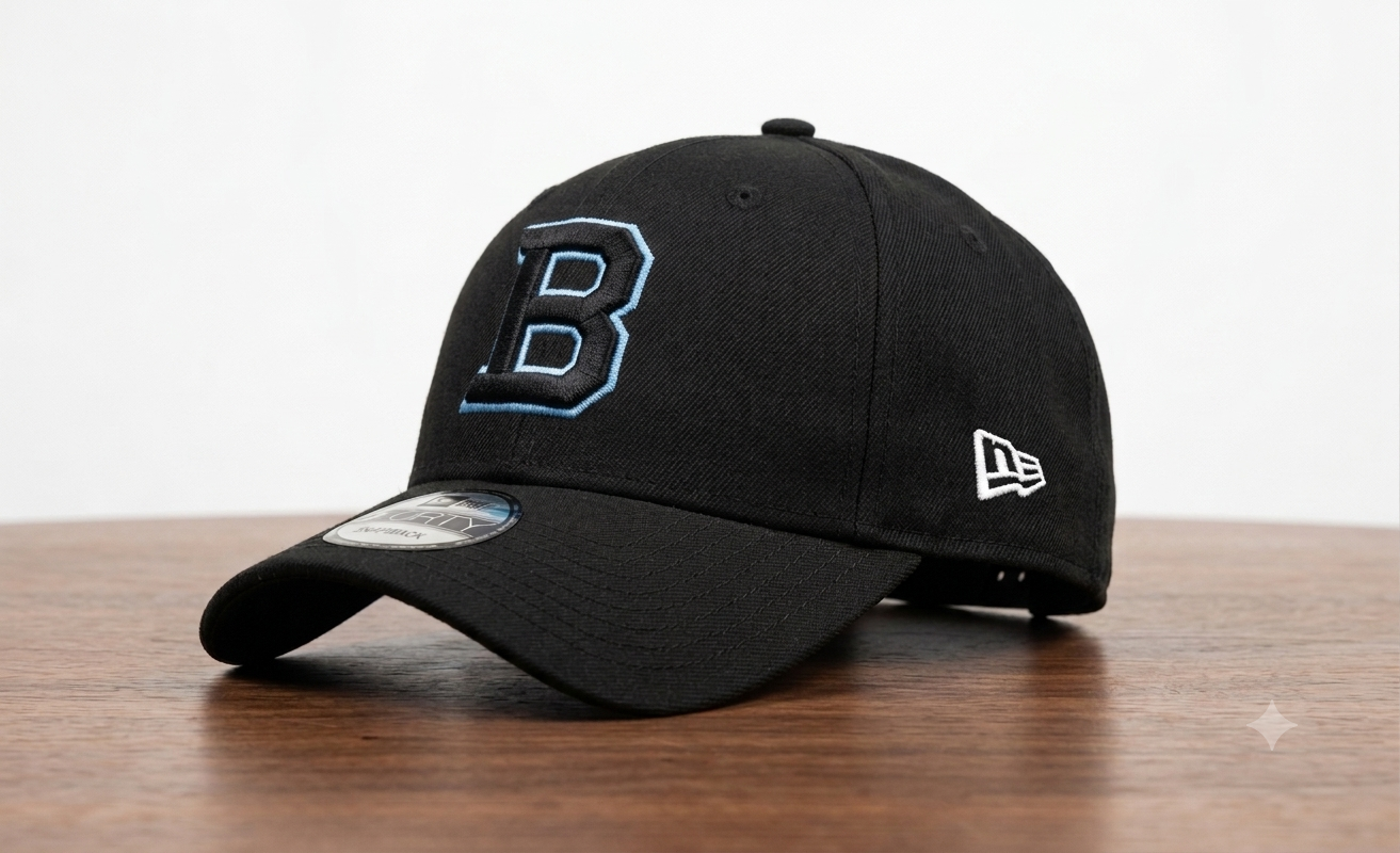 A black baseball cap with a large embroidered blue and white letter 'B' on the front, a white New Era logo on the side, and a curved brim resting on a wooden surface against a white background.