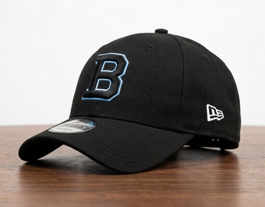 A black baseball cap with a large embroidered blue and white letter 'B' on the front, a white New Era logo on the side, and a curved brim resting on a wooden surface against a white background.