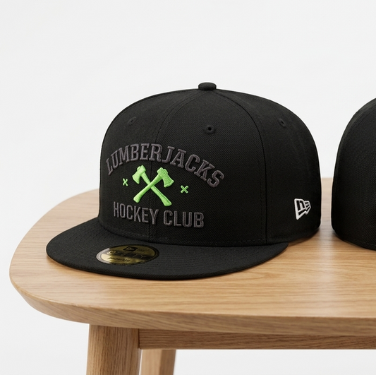 Two black fitted baseball caps displayed on a wooden table against a white background. The left cap shows the front with 'Lumberjacks Hockey Club' text and crossed axes in green and gray.