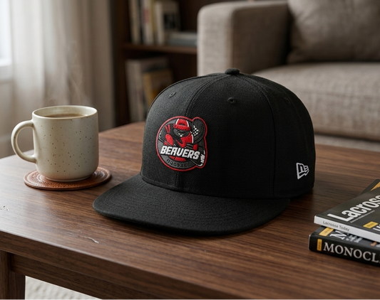 A black baseball cap with a red and white Beavers logo patch on the front, styled on a wooden table next to a ceramic mug and a magazine in a cozy living room setting.