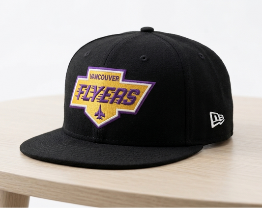 A black flat-brim baseball cap with a Vancouver Flyers patch on the front and a white New Era logo on the side, displayed on a light wood table against a white background.