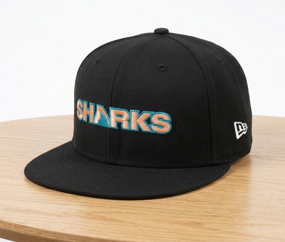 A black flat-brim baseball cap with 'SHARKS' embroidered in orange and teal across the front, resting on a light wood table against a white background. A white New Era logo is visible on the side.